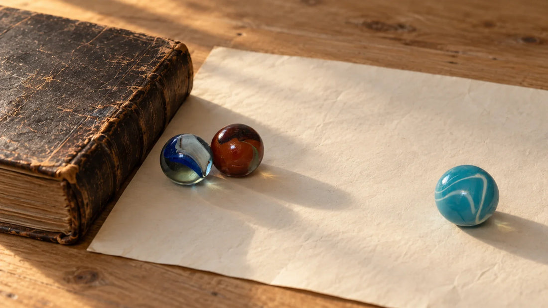 Livre ancien relié et fermé posé sur une table de bois éclairée de côté, grande feuille de papier crème à côté, deux billes de verre MesBilles posées ensemble près du livre et une troisième, bleu clair, isolée plus loin à droite